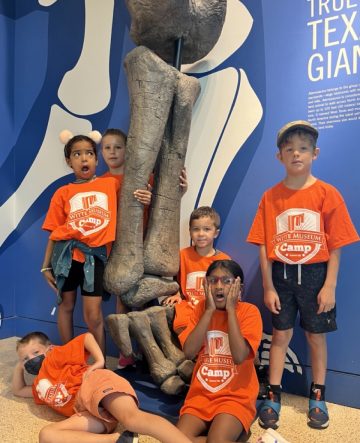 Six students in orange camp shirts, standing at the leg of a dinosaur fossil, making silly faces.