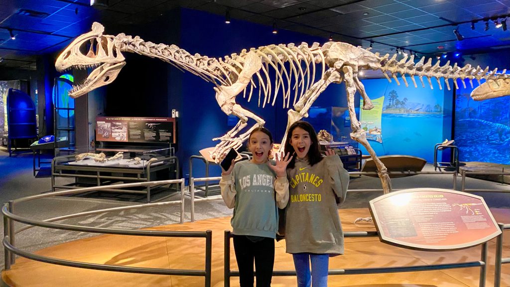 Two tween girls feigning fear with hands up and mouths open, standing in front of the cryolophosaurus model.