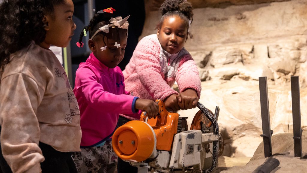 Three girls playing with an interactive, pretending to use power tools to cut into rock.