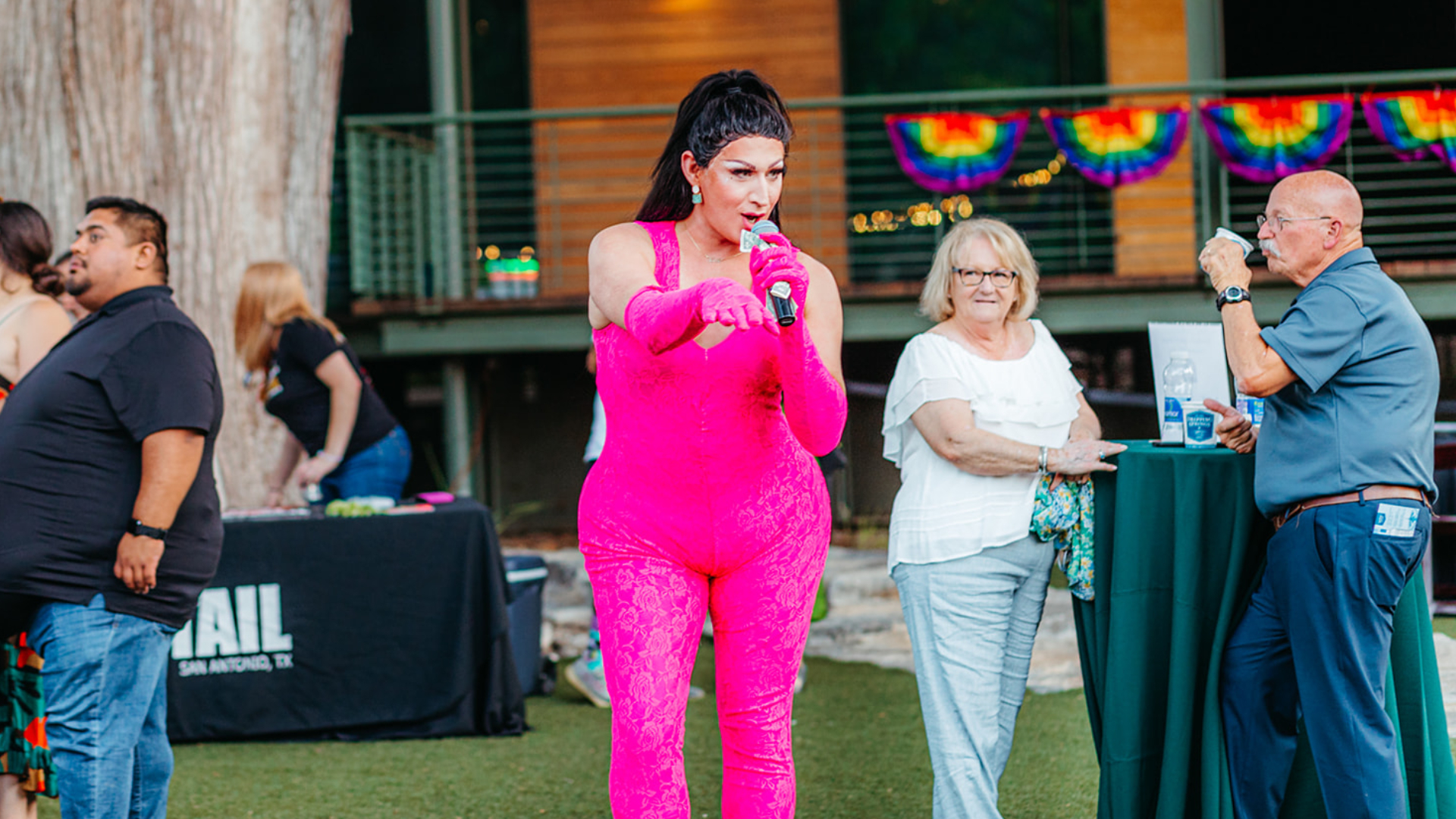 Drag queen in pink jumpsuit with high ponytail holding microphone outside on the lawn. Elderly couple stands behind, sipping cocktails. Pride flags hung along the balcony railing in the background.