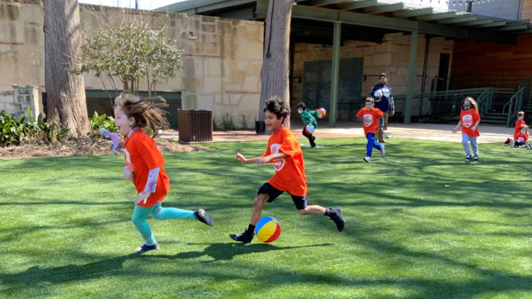 Children wearing orange camp shirts on the back lawn, running and playing what looks like tag.