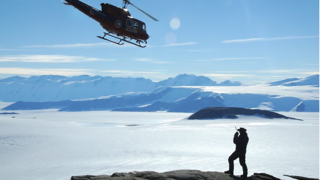 helicopter above snowy terrain. Ground is mostly flat with mountains in the distance. There is a researcher standing in the foreground on a hill talking on a walkie talkie.