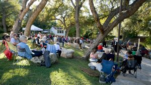 Guests sit in lawn chairs and on stone benches under the pecan trees in the amphitheater, watching musicians on stage.