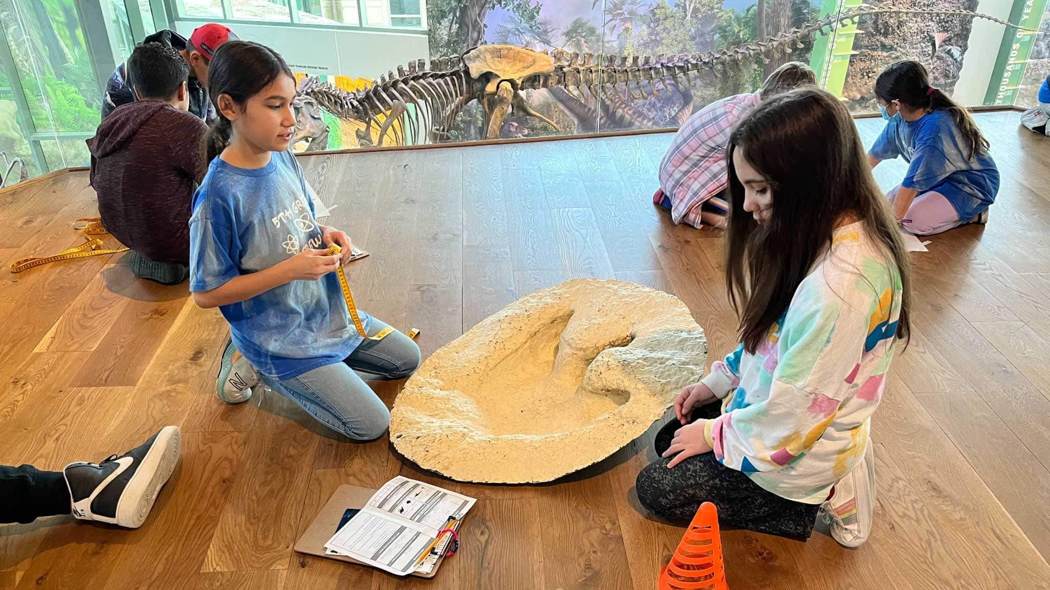 Two school-aged girls sitting on the floor next to a dinosaur track cast.