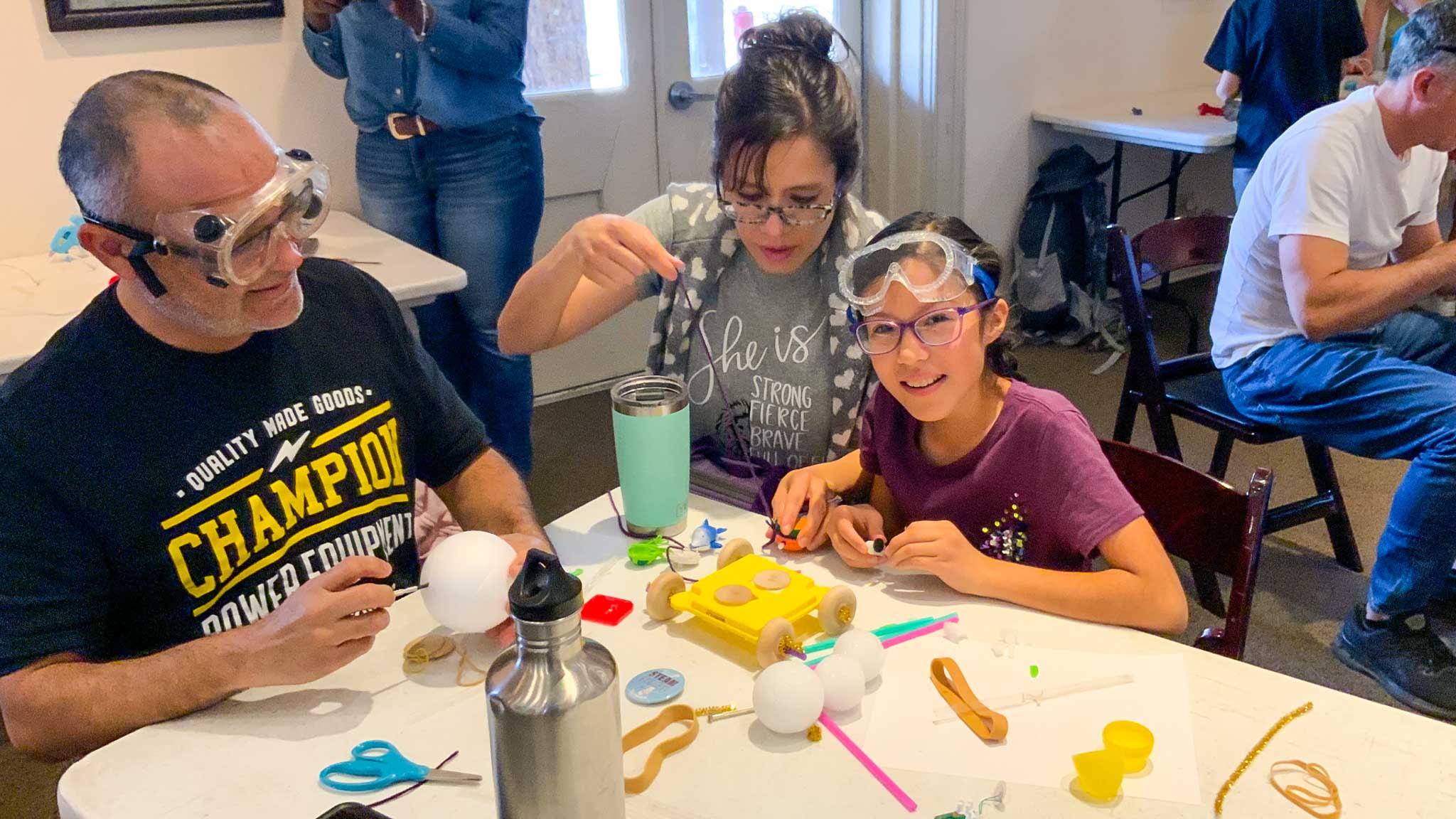Two parents and young girl wearing goggles, smiling and sitting at a table covered in craft materials.