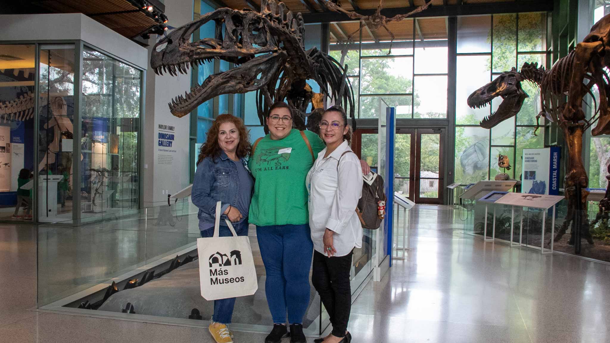 3 female educators standing in front of a dinosaur fossil, smiling. One holds a "mas museos" tote bag.