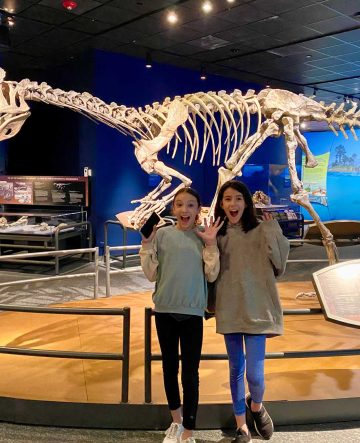 Two tween girls feigning fear with hands up and mouths open, standing in front of the cryolophosaurus model.