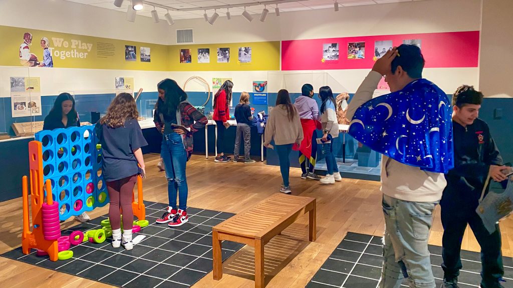 Teens and tweens playing in the exhibition. Some are putting on costumes, some are playing giant connect four, some are looking at the display cases.