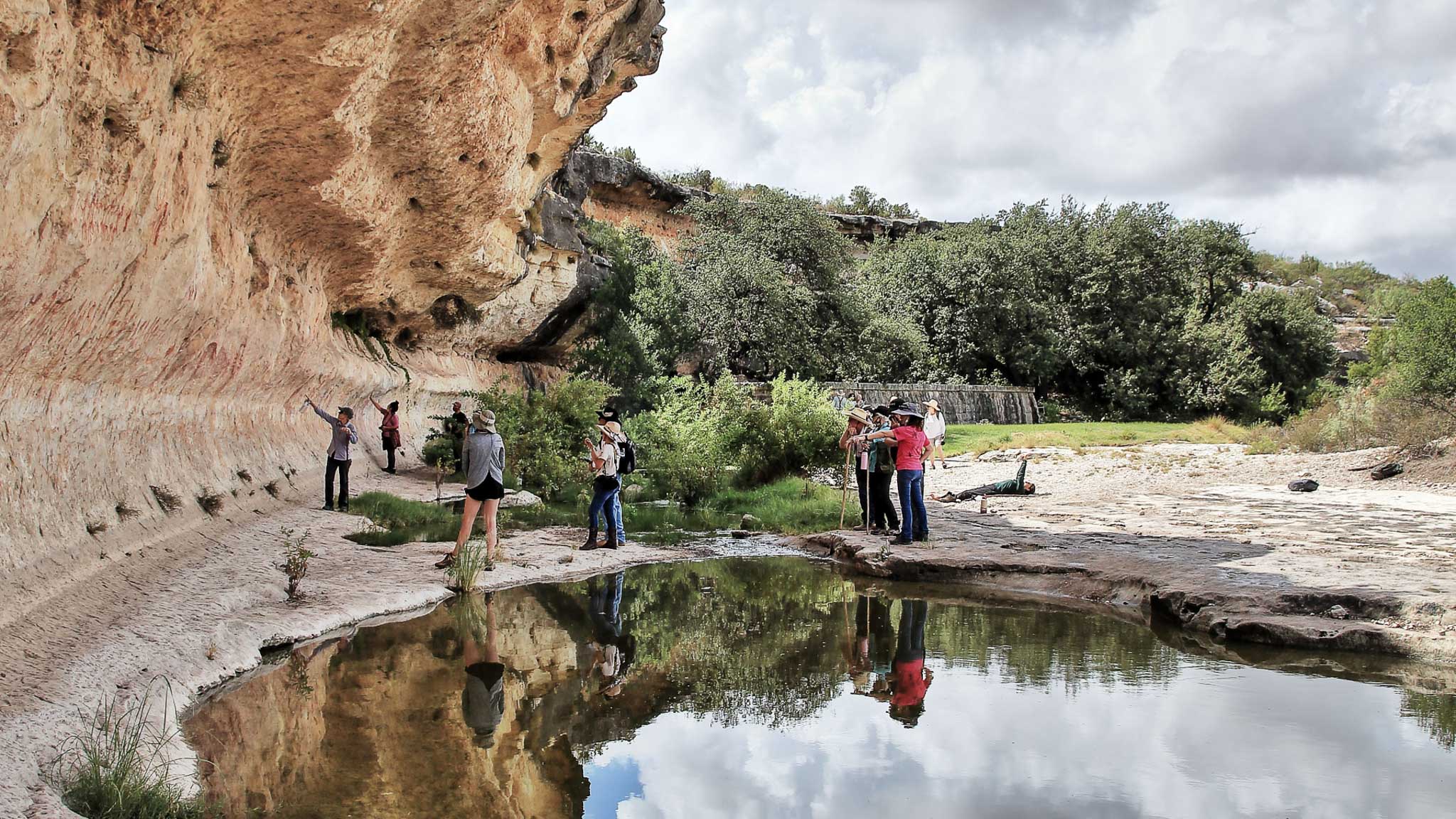 A spring in the foreground, with people standing on the other side, looking at the cliff with rock art.