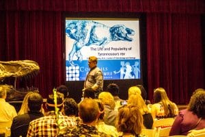 Dr. Thomas Adams standing in front of a crowd with a screen behind him reading "the life and popularity of tyrannosaurus rex."