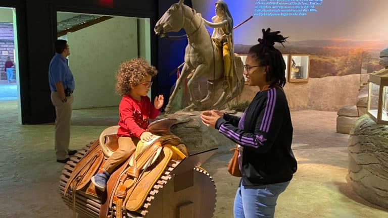 mother stands near child on the cowboy saddle.