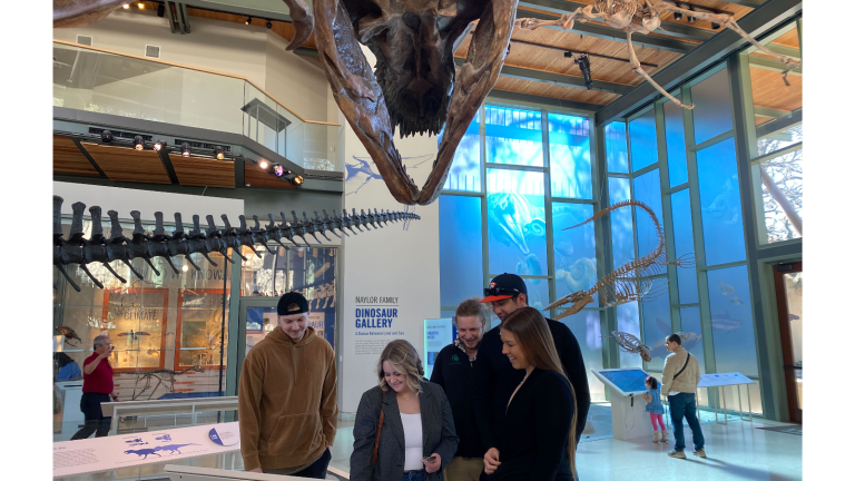 young adults stand under t. rex and look at screen display.