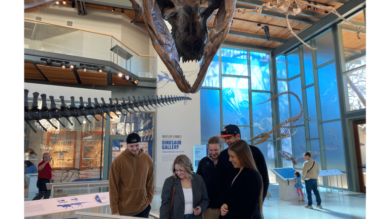 young adults stand under t. rex and look at screen display.