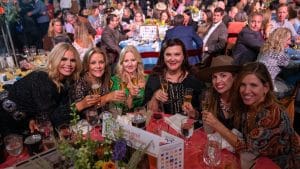 Six women gather around a table, holding sparkling wine and smiling.