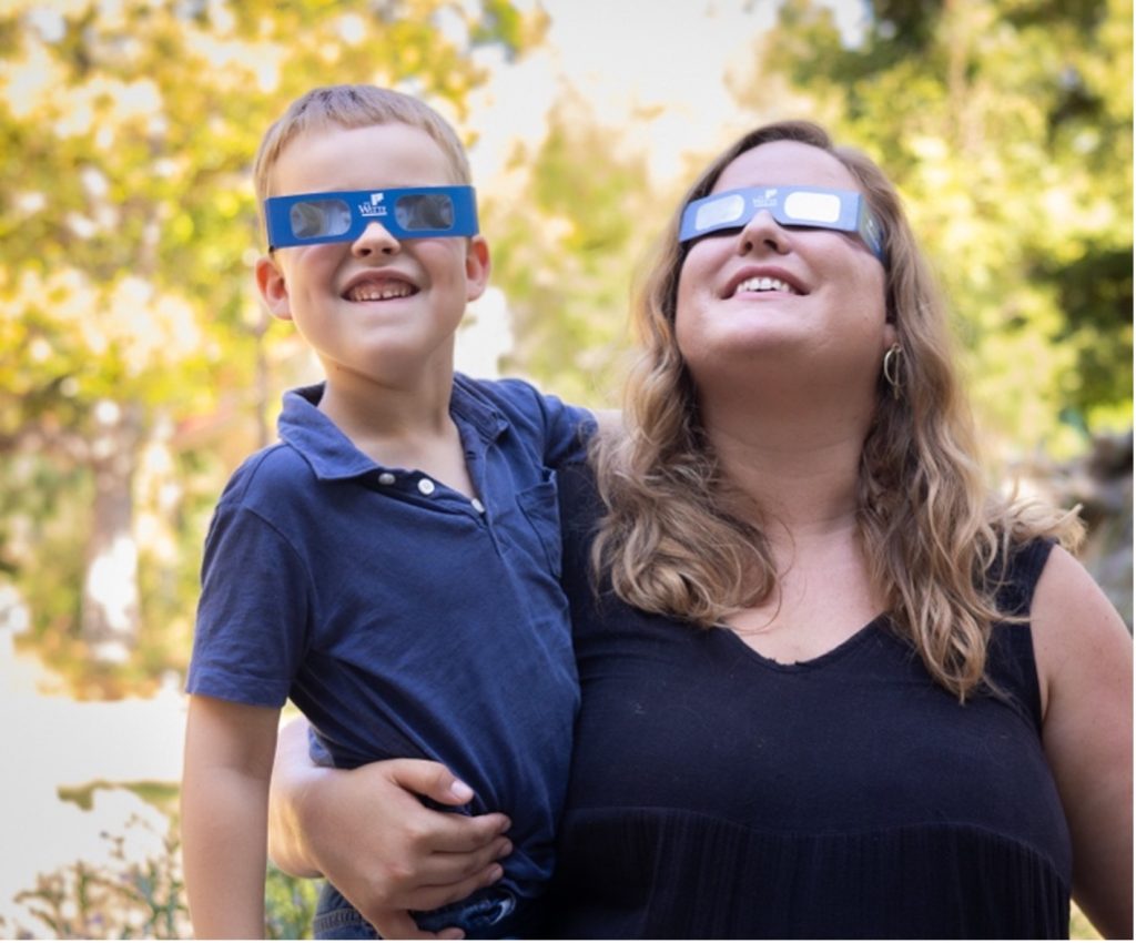 Mother and son wear eclipse glasses while looking at the sky.