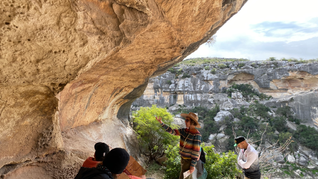 guide speaking and gesturing to rock art. Hikers stand and sit around.