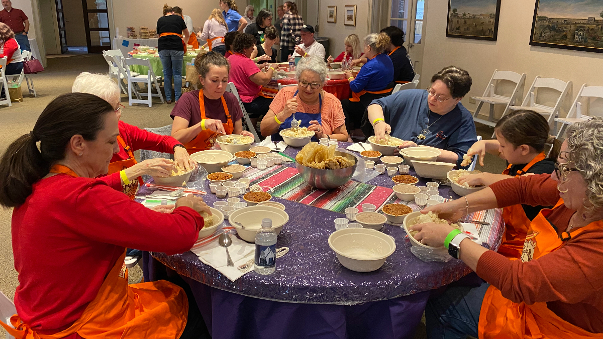 7 people sit around a table making tamales.