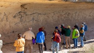 group gathers around a rock art drawing of a horse on cave wall.