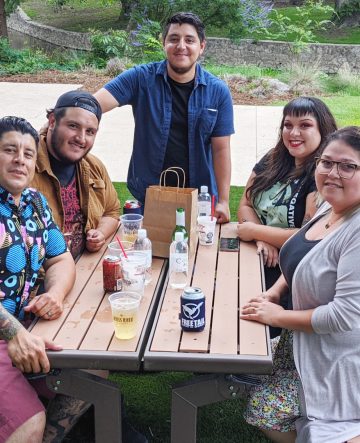 group of 5 smiles and sips beers while sitting at a picnic bench.