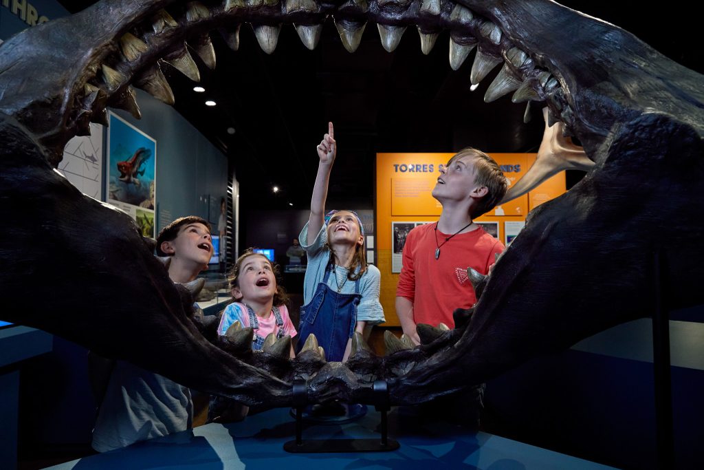 group of children stand inside shark jaws and point at teeth.